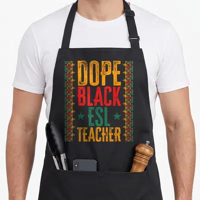 Second english chef apron - Stacked DOPE BLACK ESL TEACHER text in orange, red, green with geometric borders and two — shown on a black cotton canvas apron against a white background