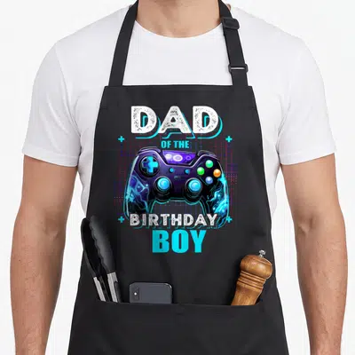 Retro birthday video game apron - DAD OF THE BIRTHDAY BOY text surrounding a neon purple and cyan game controller — shown on a black cotton canvas apron against a white background