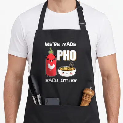 Each vietnamese pho apron - We're Made Pho Each Other pun with cartoon sriracha bottle and smiling noodle bowl — shown on a black cotton canvas apron against a white background