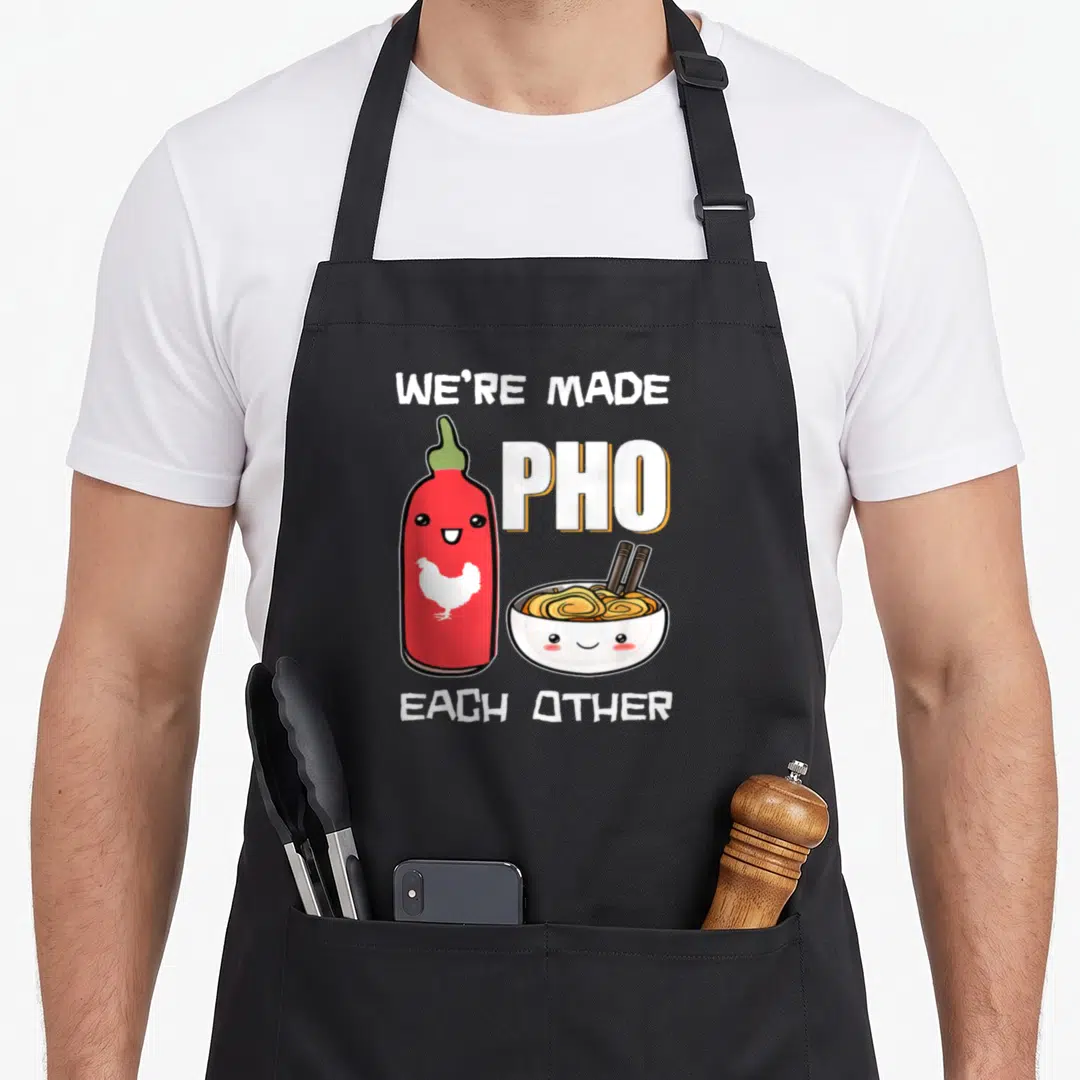 Each vietnamese pho apron - We're Made Pho Each Other pun with cartoon sriracha bottle and smiling noodle bowl — shown on a black cotton canvas apron against a white background
