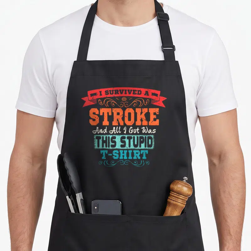 Funny survivor stroke awareness apron - Distressed text reading I survived a stroke and all I got was this stupid — shown on a black cotton canvas apron against a white background