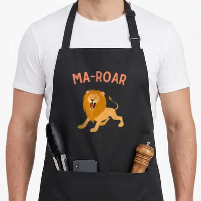Plate passover apron - Orange Ma-Roar text arched over a cartoon roaring lion — shown on a black cotton canvas apron against a white background