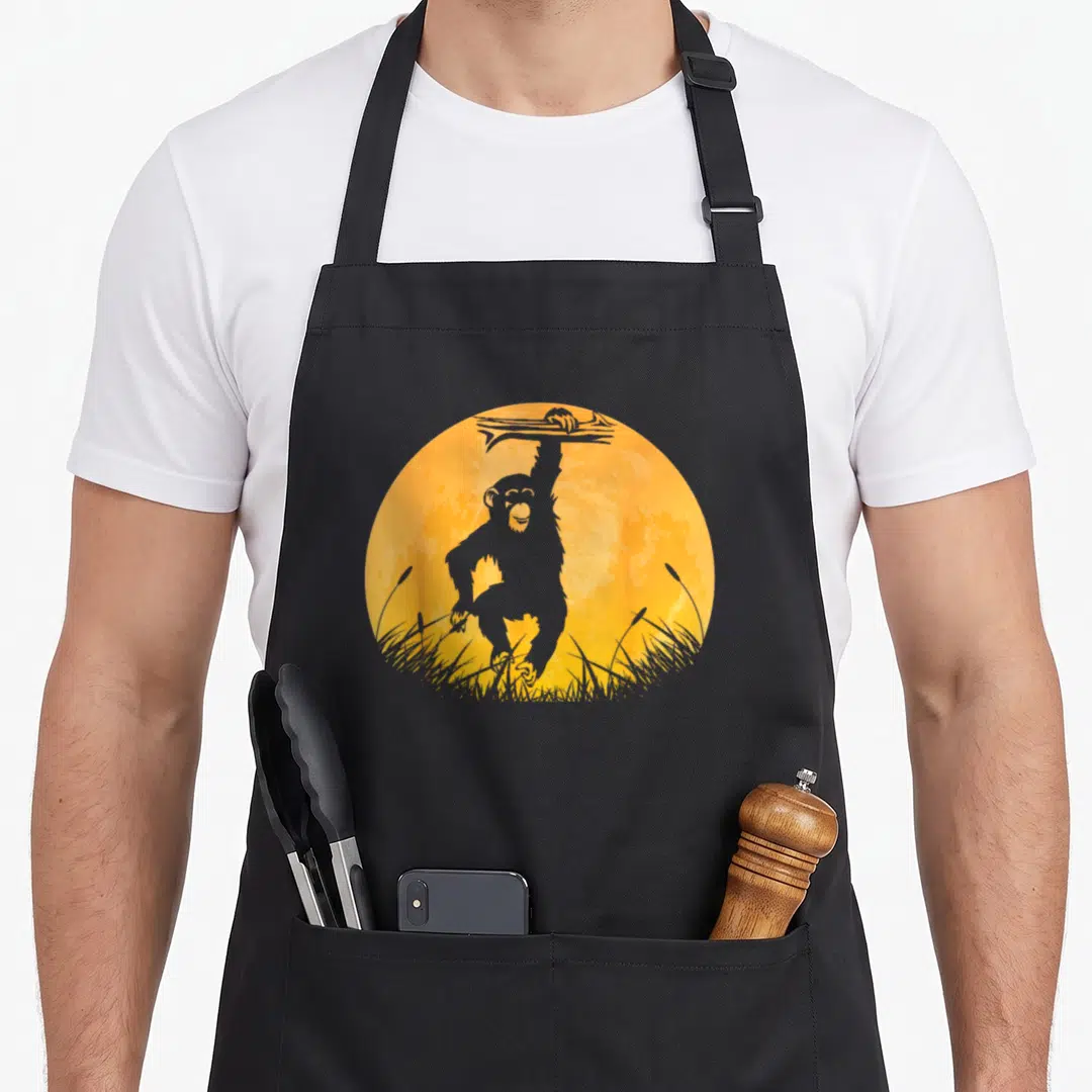 Chimpanzee halloween apron - Grey cartoon chimpanzee hanging from a branch inside an orange circle above grass — shown on a black cotton canvas apron against a white background