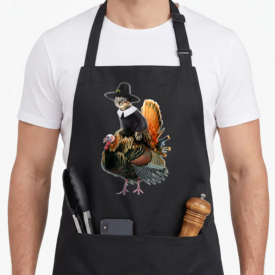 Cat pilgrim apron - Photo of a tabby cat in a black pilgrim hat riding a large turkey with spread tail feathers — shown on a black cotton canvas apron against a white background