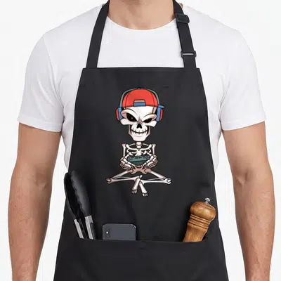 Skull video halloween apron - Cartoon skeleton in red cap and blue headphones holding a game controller while sitting — shown on a black cotton canvas apron against a white background