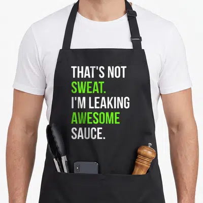 Leaking funny sauce apron - Stacked white and lime green text reading THAT'S NOT SWEAT I'M LEAKING AWESOME SAUCE — shown on a black cotton canvas apron against a white background