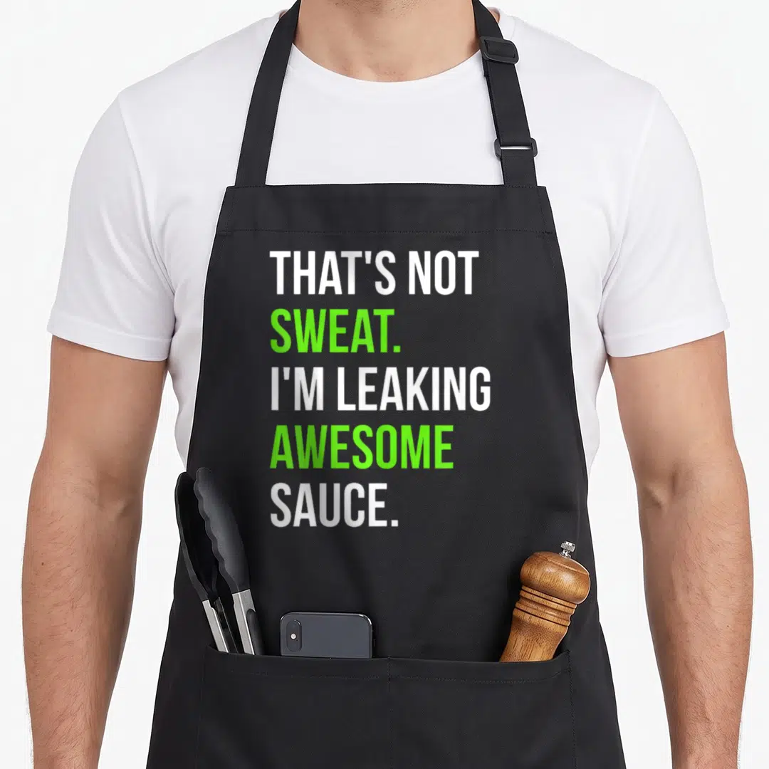 Leaking funny sauce apron - Stacked white and lime green text reading THAT'S NOT SWEAT I'M LEAKING AWESOME SAUCE — shown on a black cotton canvas apron against a white background