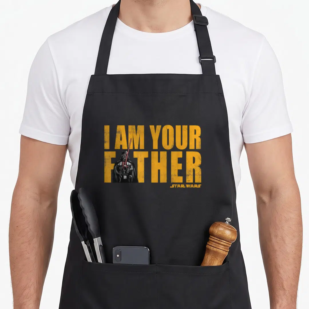 Vintage father falcon dad apron - I AM YOUR FATHER text with Darth Vader figure in yellow and black distressed style — shown on a black cotton canvas apron against a white background