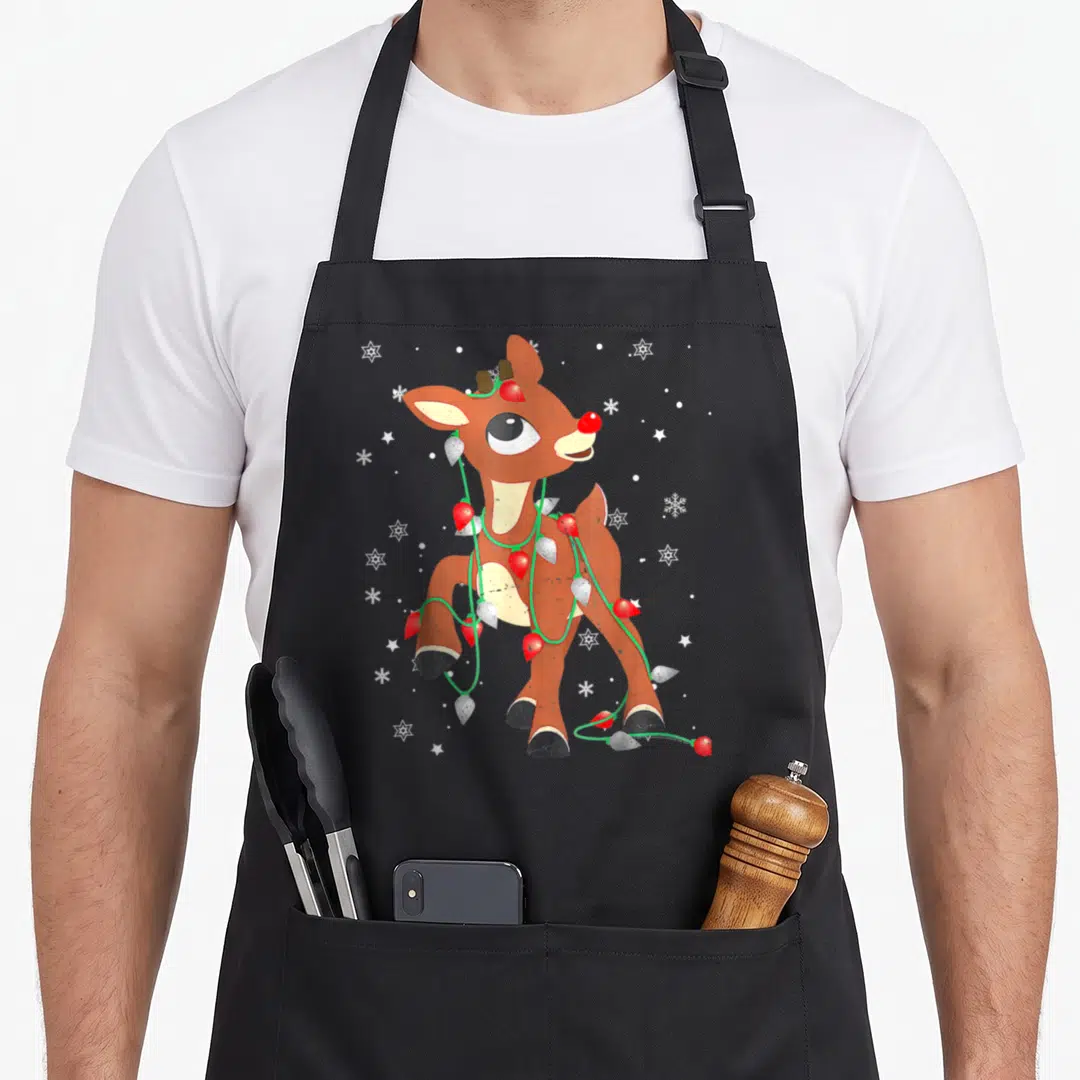 Rudolph red christmas apron - Cartoon brown reindeer wrapped in red and white lights with white snowflakes — shown on a black cotton canvas apron against a white background