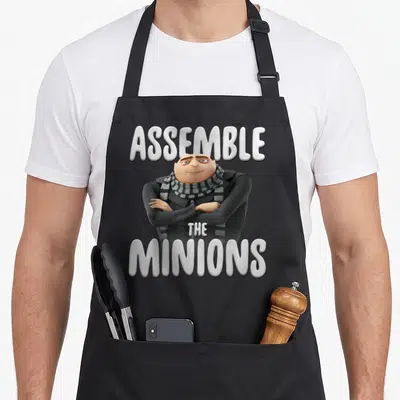 Assemble general apron - Gru with arms crossed and text Assemble The Minions in white and grey — shown on a black cotton canvas apron against a white background