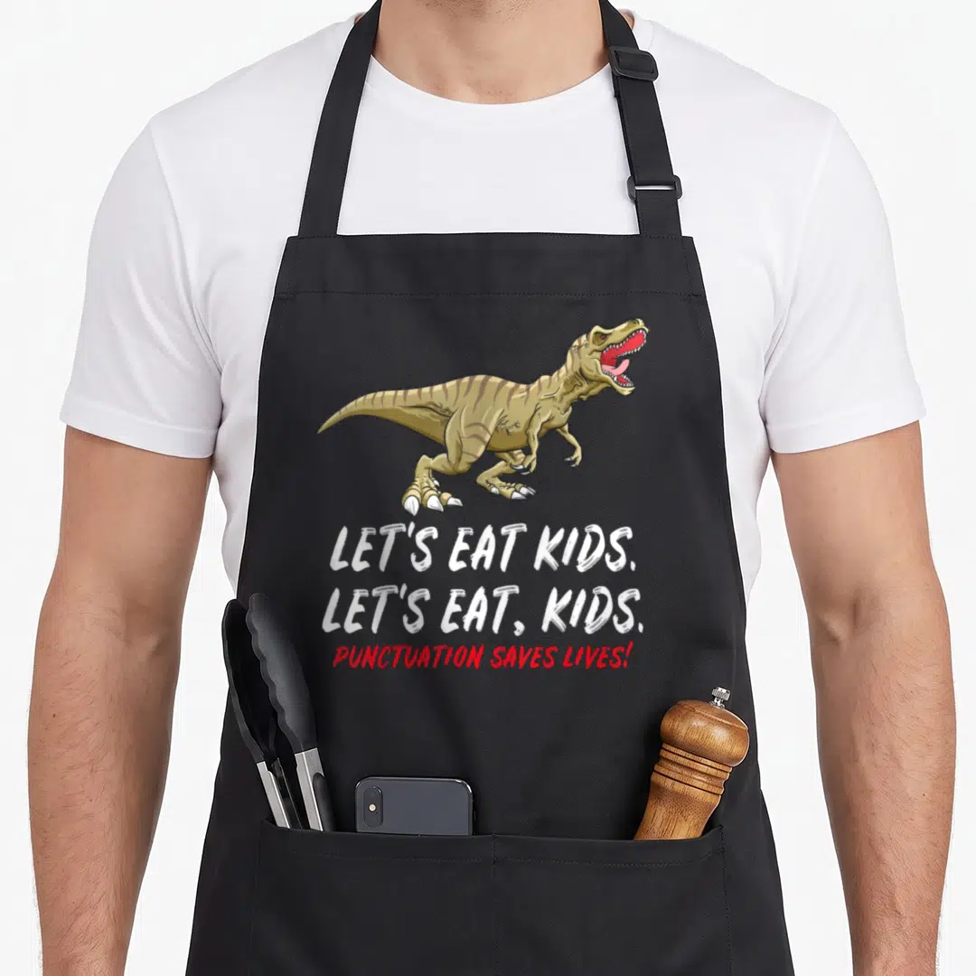 School let's eat apron - Brown T-Rex above white and red text reading Let's Eat Kids Punctuation Saves Lives — shown on a black cotton canvas apron against a white background