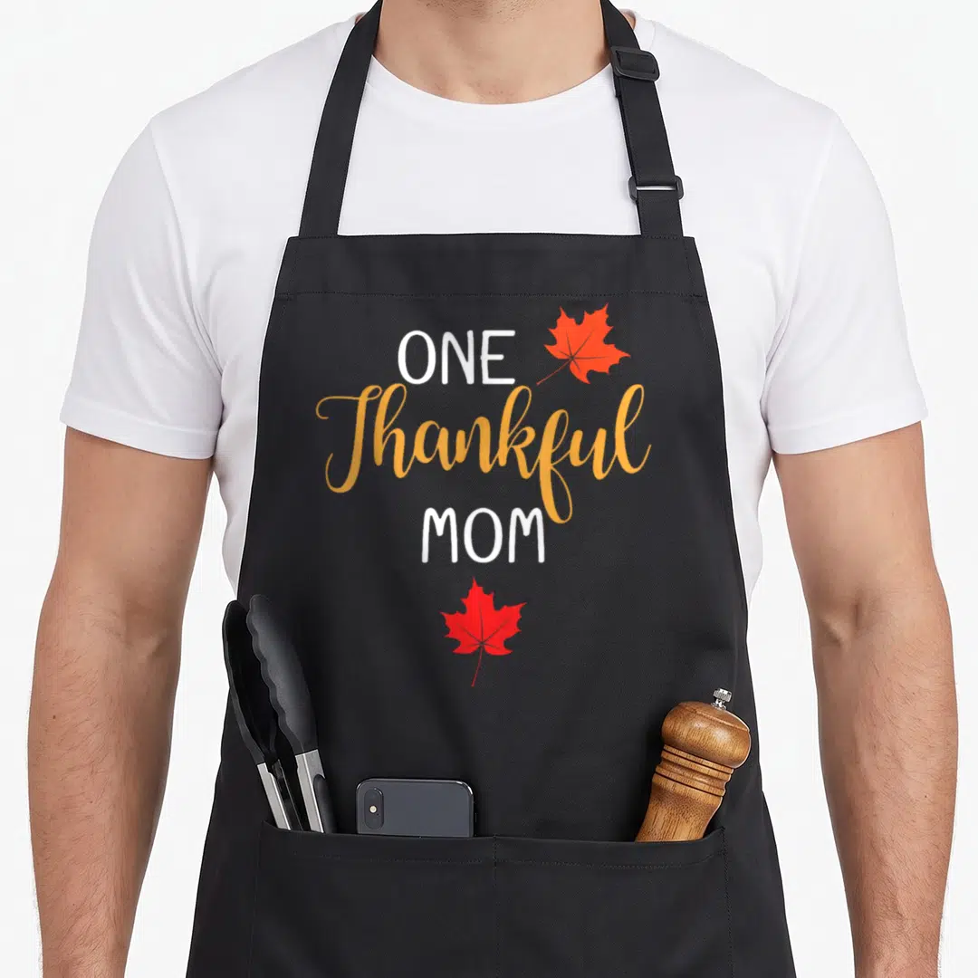 Cute thanksgiving thankful apron - Stacked text One Thankful Mom with orange and red maple leaves — shown on a black cotton canvas apron against a white background