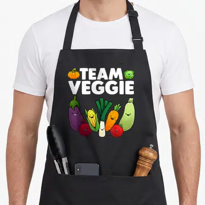 Plant veggie apron - TEAM VEGGIE text with pumpkin, cabbage, and seven smiling cartoon vegetables in color — shown on a black cotton canvas apron against a white background