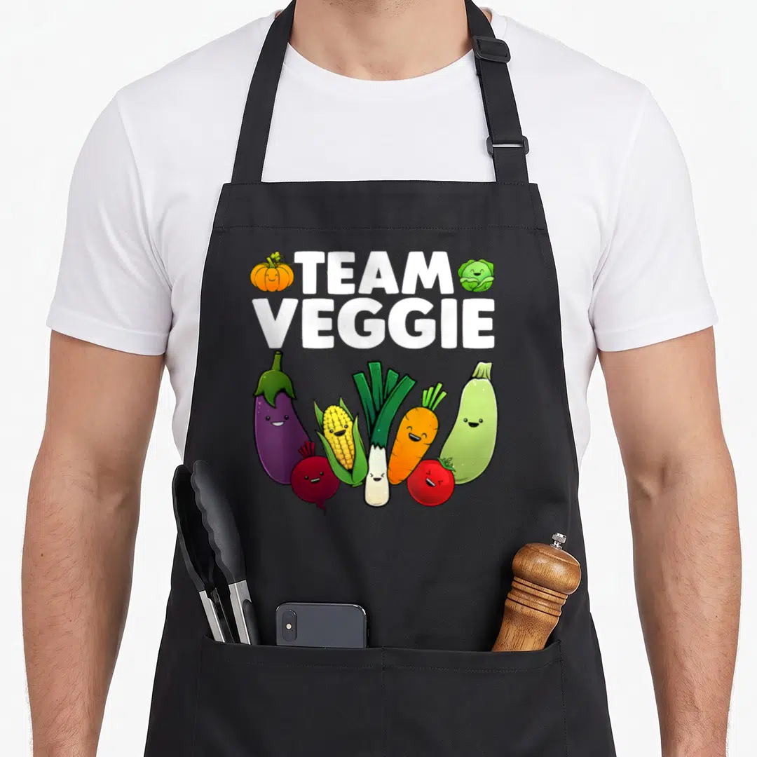 Plant veggie apron - TEAM VEGGIE text with pumpkin, cabbage, and seven smiling cartoon vegetables in color — shown on a black cotton canvas apron against a white background