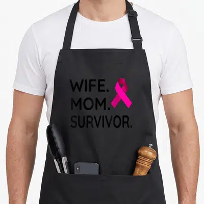 Wife survivor breast cancer awareness aprons - WIFE. MOM. SURVIVOR. text next to a pink awareness ribbon on black fabric — shown on a black cotton canvas apron against a white background