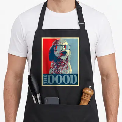 Cute dood goldendoodle apron - Retro dog portrait with sunglasses above THE DOOD text on split red and blue background — shown on a black cotton canvas apron against a white background