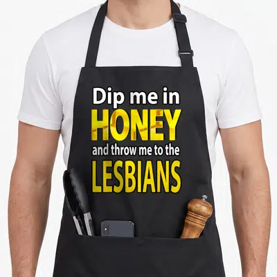 Dip throw apron - Stacked yellow and white text reading Dip me in HONEY and throw me to the LESBIANS with black outlines — shown on a black cotton canvas apron against a white background