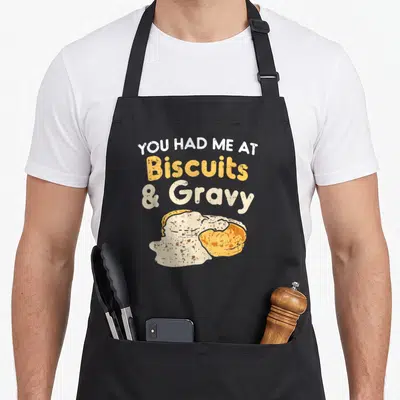 Biscuit gravy food aprons - Stacked text reading You Had Me At Biscuits And Gravy above a distressed illustration — shown on a black cotton canvas apron against a white background