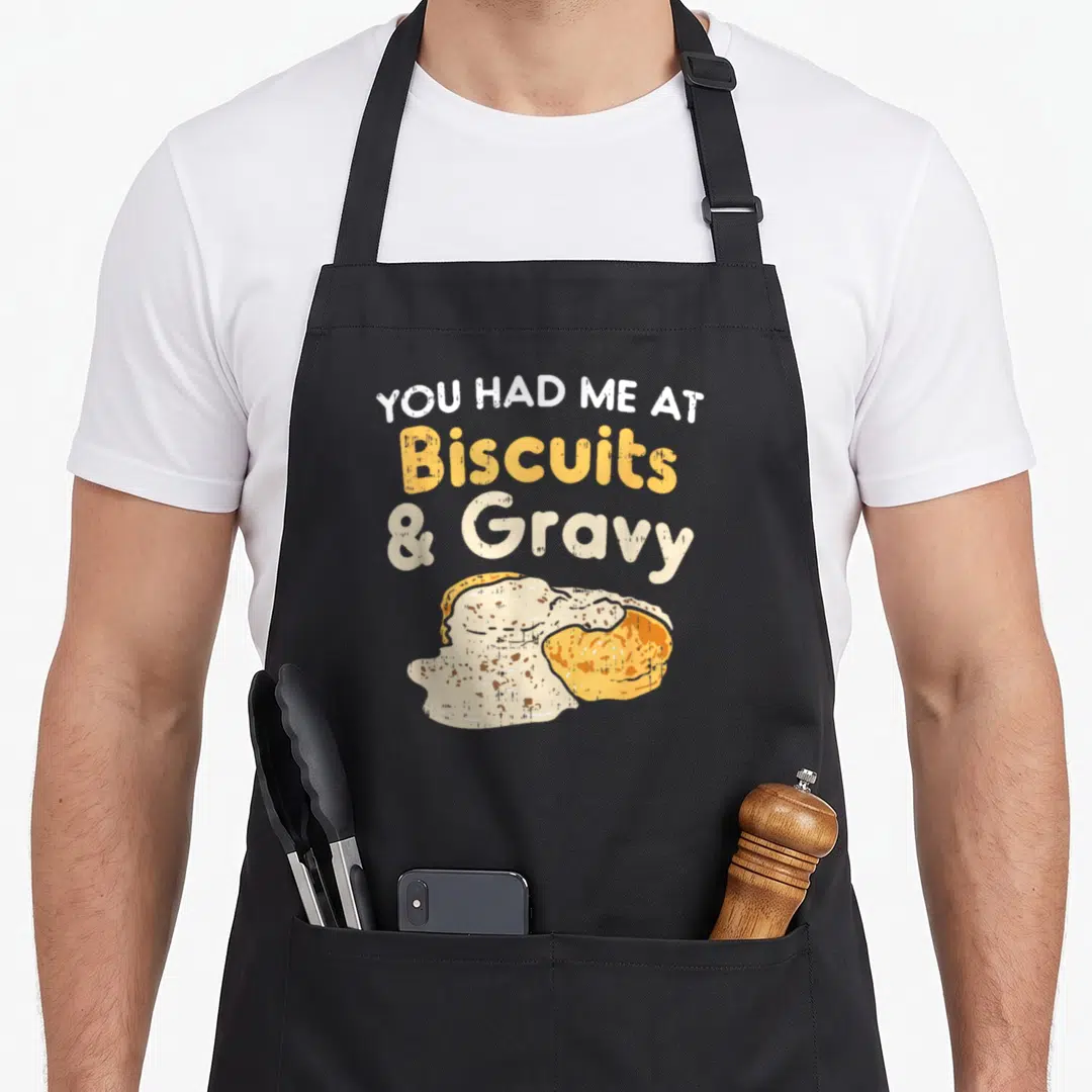 Biscuit gravy food aprons - Stacked text reading You Had Me At Biscuits And Gravy above a distressed illustration — shown on a black cotton canvas apron against a white background