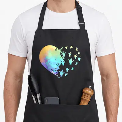 Hand teacher apron - Watercolor heart dissolving into rainbow colored I love you sign language hand gestures — shown on a black cotton canvas apron against a white background