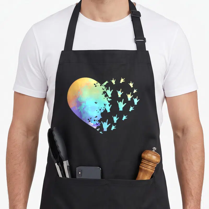 Hand teacher apron - Watercolor heart dissolving into rainbow colored I love you sign language hand gestures — shown on a black cotton canvas apron against a white background