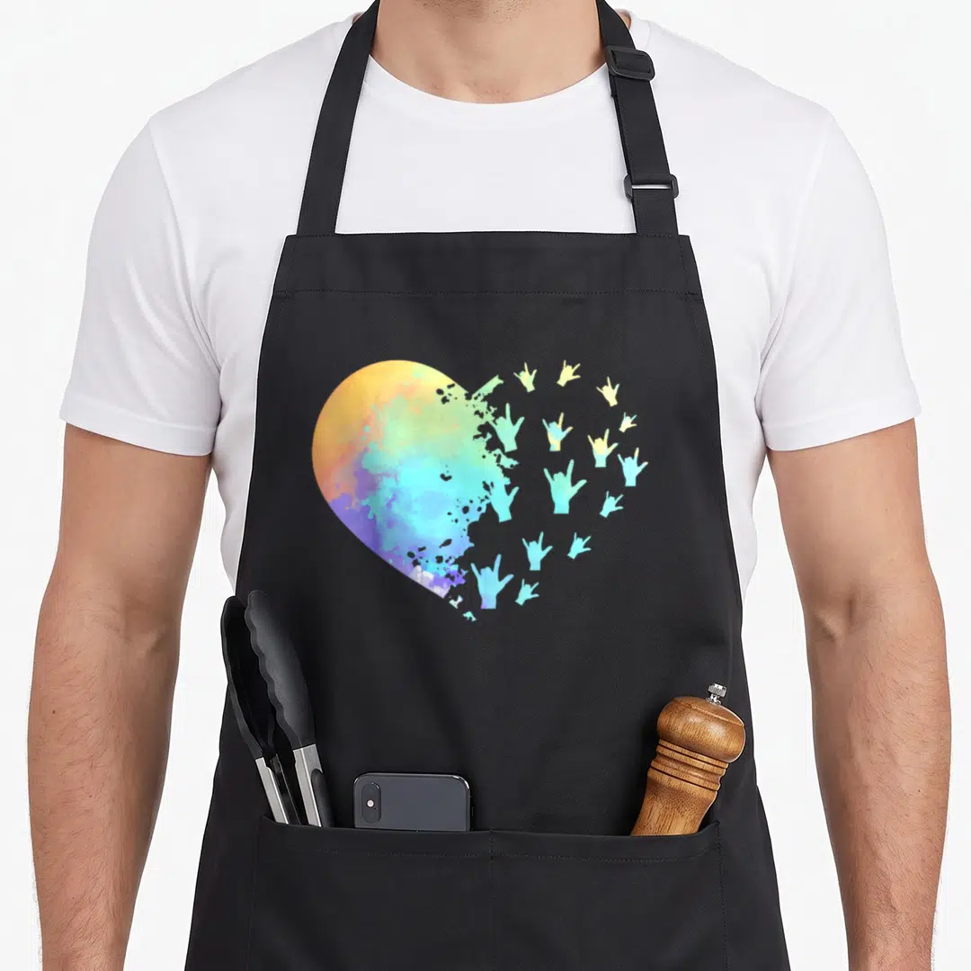 Hand teacher apron - Watercolor heart dissolving into rainbow colored I love you sign language hand gestures — shown on a black cotton canvas apron against a white background