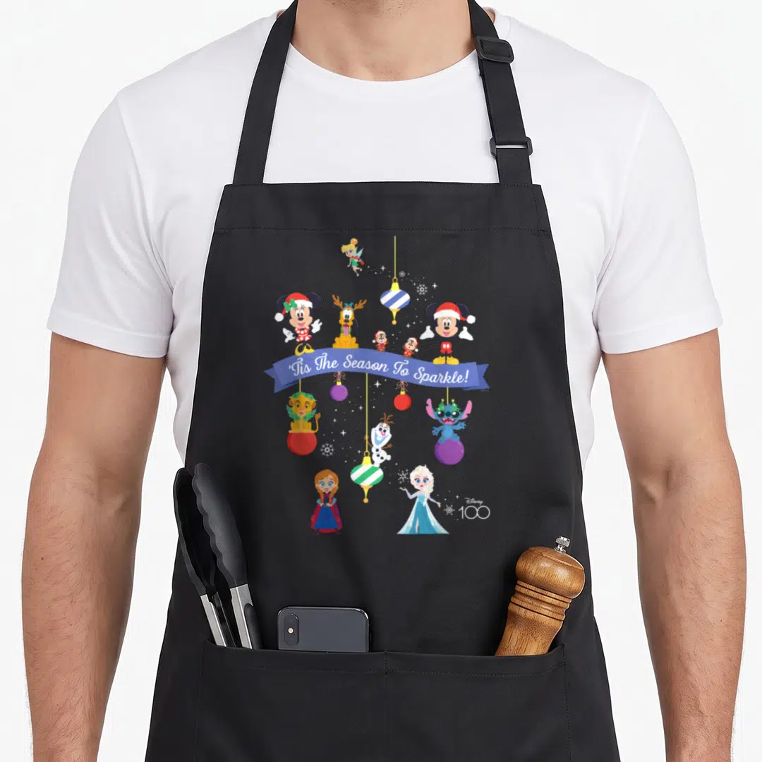 Tis christmas apron - Disney characters on blue ribbon with ornaments and snowflakes celebrating centennial — shown on a black cotton canvas apron against a white background