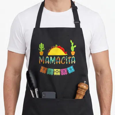 Cactus mexican apron - MAMACITA text below yellow taco, two cacti, five colorful papel picado banners — shown on a black cotton canvas apron against a white background