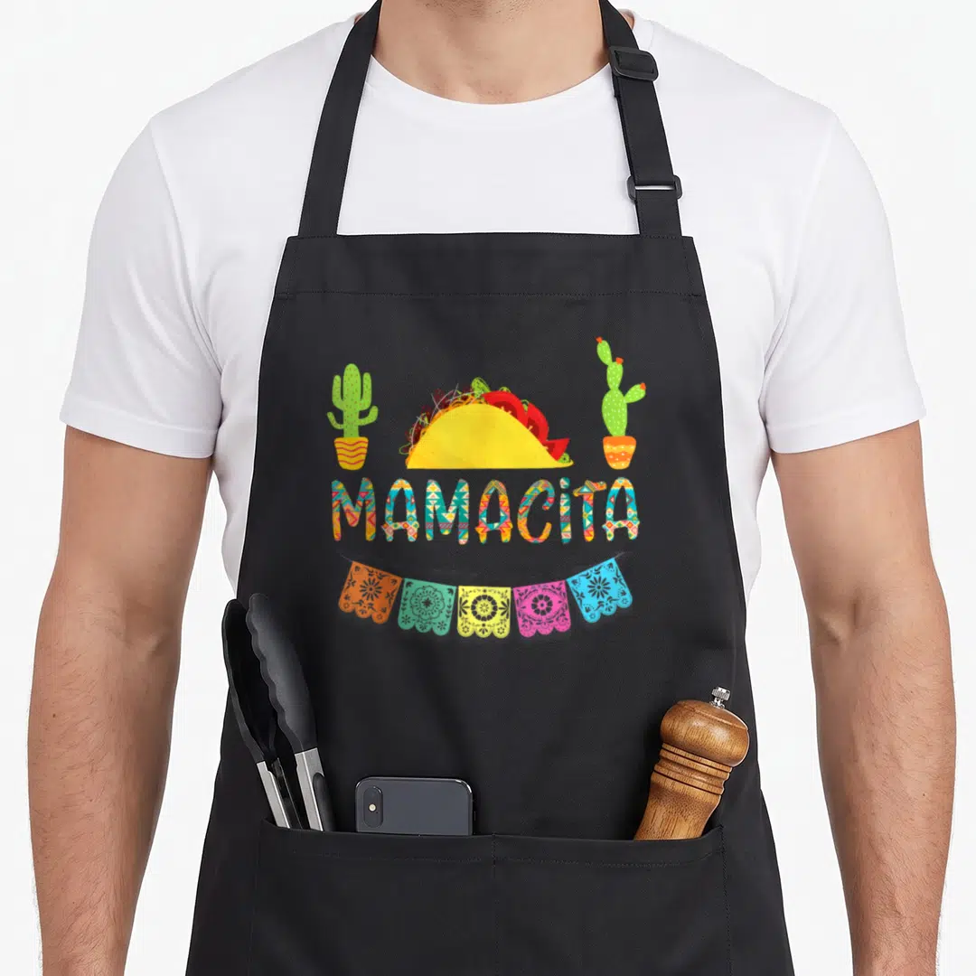 Cactus mexican apron - MAMACITA text below yellow taco, two cacti, five colorful papel picado banners — shown on a black cotton canvas apron against a white background
