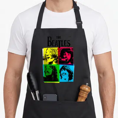 Cmyk beatle apron - THE Beatles text above four pop art portraits on yellow red green and blue backgrounds — shown on a black cotton canvas apron against a white background