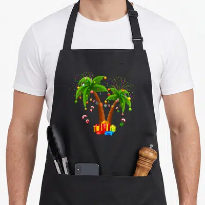 Tropical christmas tree apron - Two decorated palm trees with presents, parachutes, and fireworks in green red yellow — shown on a black cotton canvas apron against a white background