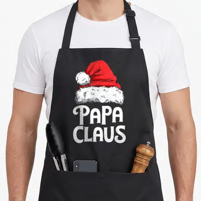 Papa santa claus apron - Red and white Santa hat above stacked white PAPA CLAUS text in cartoon style — shown on a black cotton canvas apron against a white background