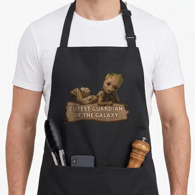 Studio galaxy apron - Baby Groot lying on wooden sign reading cutest guardian of the galaxy — shown on a black cotton canvas apron against a white background