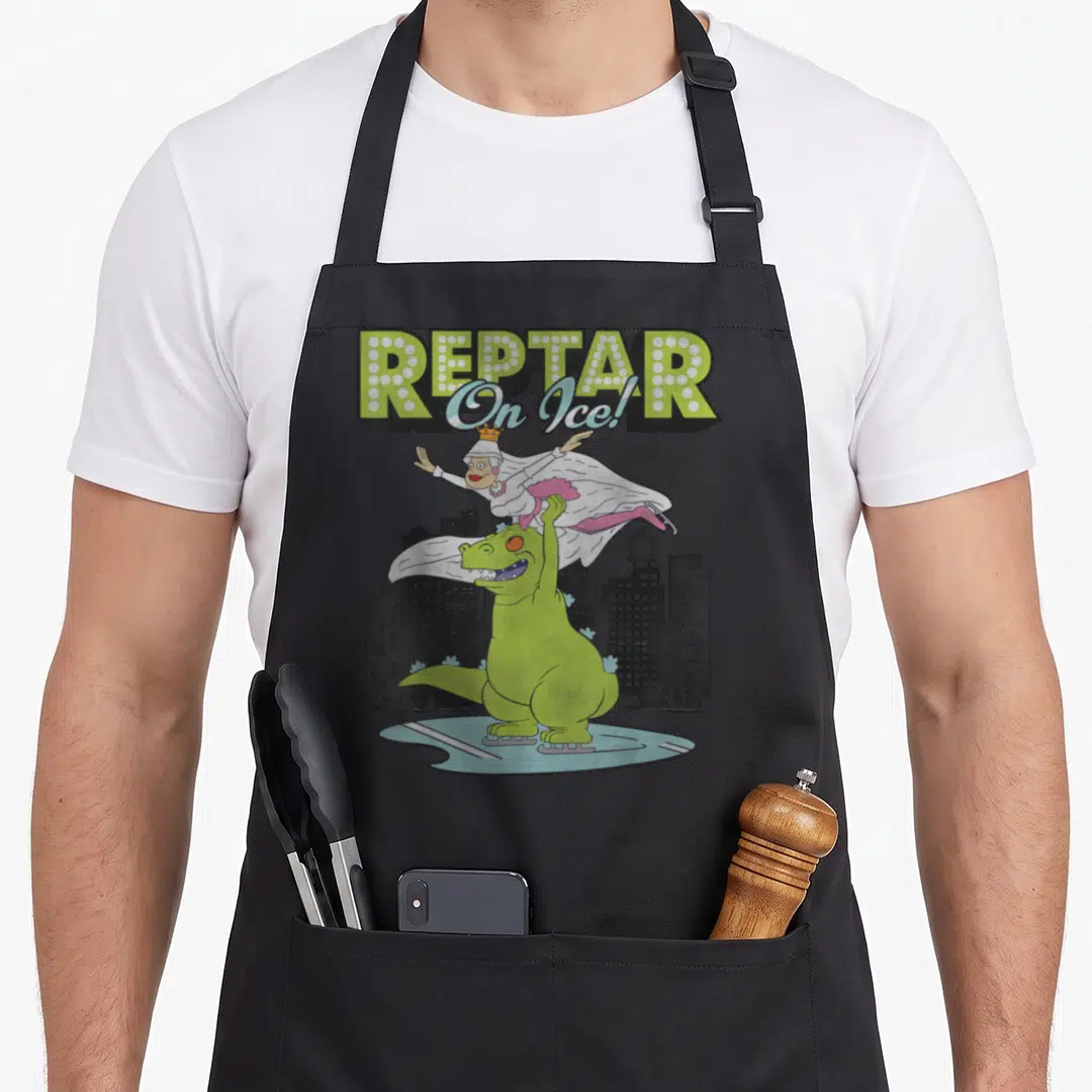 Reptar frappe apron - Green dinosaur on ice skates lifting a crowned woman under REPTAR text — shown on a black cotton canvas apron against a white background