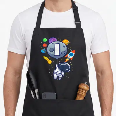 Vintage astronaut birthday apron - Cartoon astronaut holding moon balloon with planets and rocket in space — shown on a black cotton canvas apron against a white background