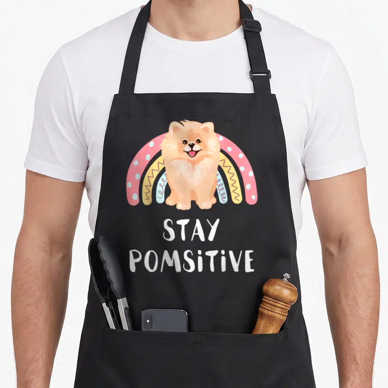 Pom dog mom apron - Cartoon Pomeranian sitting before a multi-colored rainbow with Stay Pomsitive text below — shown on a black cotton canvas apron against a white background