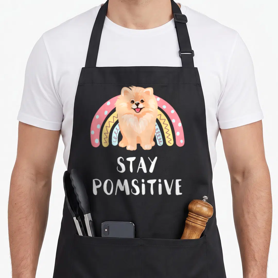 Pom dog mom apron - Cartoon Pomeranian sitting before a multi-colored rainbow with Stay Pomsitive text below — shown on a black cotton canvas apron against a white background