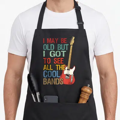 May concert apron - Red electric guitar standing vertically beside stacked distressed text reading I May Be Old But I — shown on a black cotton canvas apron against a white background