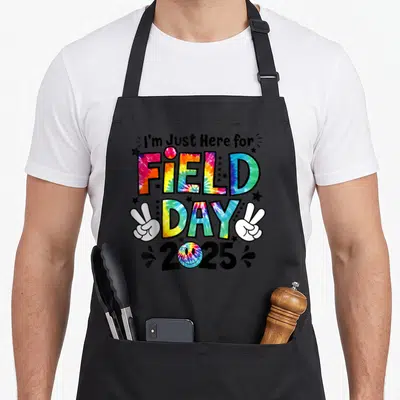 Field tie-dye apron - I'm Just Here for Field Day 2025 tie-dye text with peace signs and smiley face — shown on a black cotton canvas apron against a white background