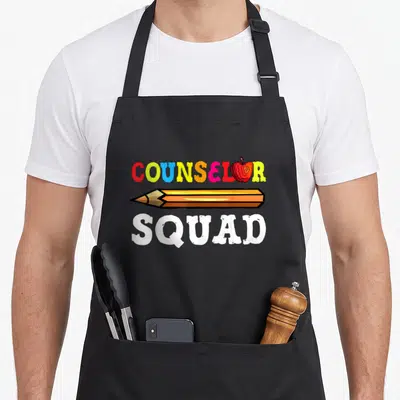 Counselor squad school teacher apron - COUNSELOR SQUAD text with apple replacing O and large pencil in cartoon colors — shown on a black cotton canvas apron against a white background