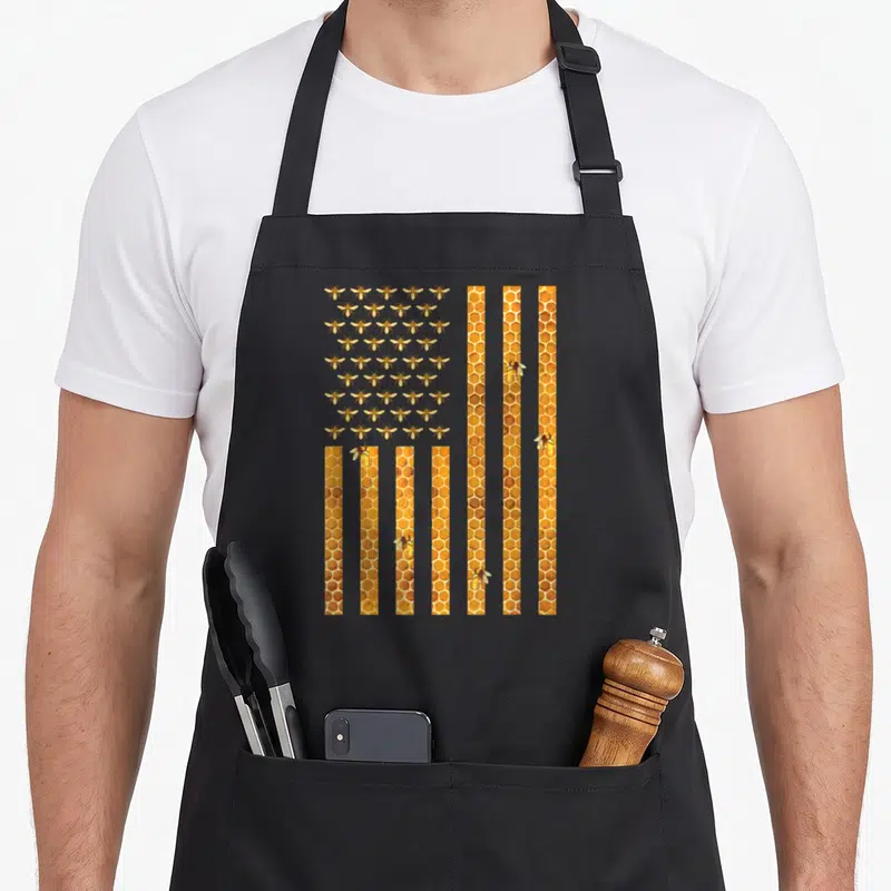 Bee american flag apron - Stylized flag with rows of bees in canton and vertical honeycomb stripes in yellow orange — shown on a black cotton canvas apron against a white background