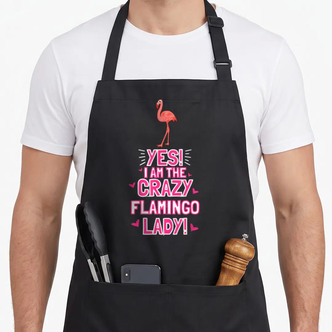 Outfit flamingo apron - Pink flamingo illustration above stacked Yes I Am The Crazy Flamingo Lady text with small hearts — shown on a black cotton canvas apron against a white background
