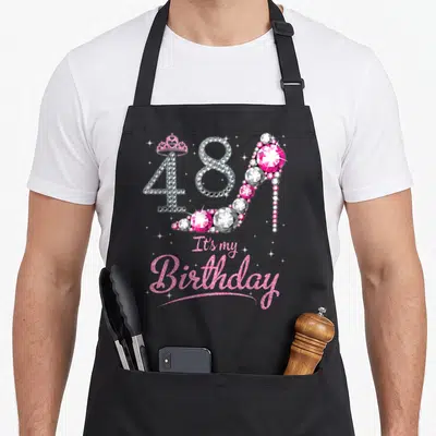 Queen nana aprons - Jeweled 48 high heel shoe with crown and It's my Birthday text in pink and silver — shown on a black cotton canvas apron against a white background