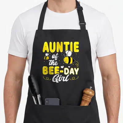 Auntie bee aprons - AUNTIE of the BEE-DAY Girl text with two cartoon bees, hearts, and dots — shown on a black cotton canvas apron against a white background