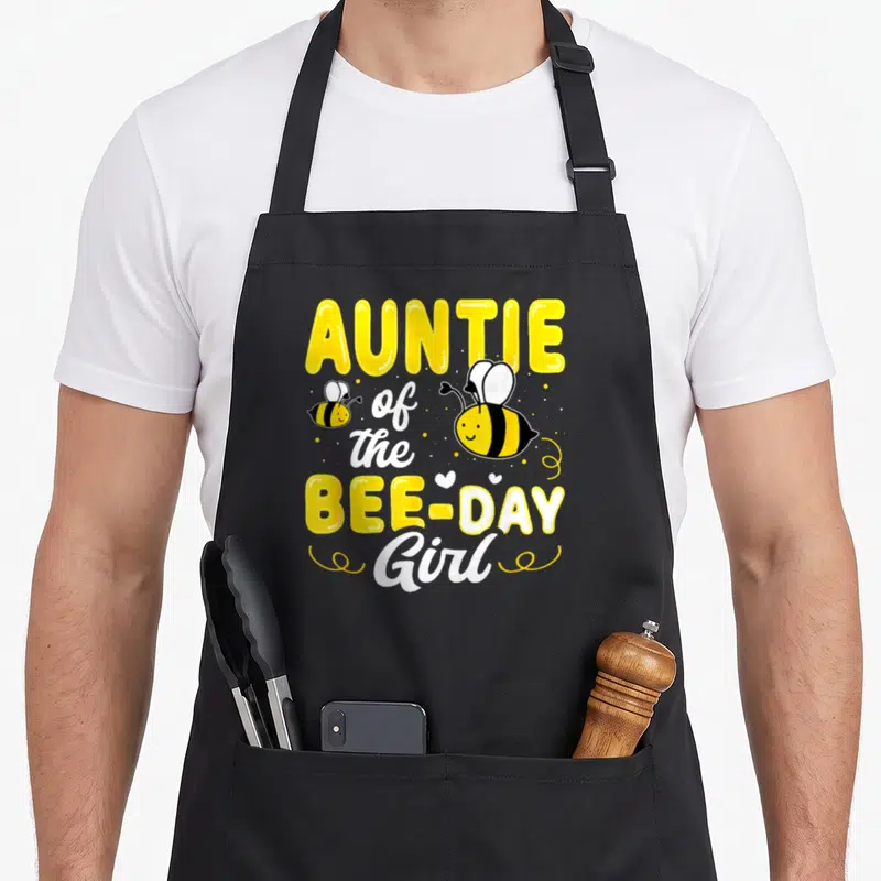 Auntie bee aprons - AUNTIE of the BEE-DAY Girl text with two cartoon bees, hearts, and dots — shown on a black cotton canvas apron against a white background