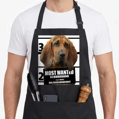Wanted dog apron - Bloodhound head holding a black Most Wanted sign with case number 1587552 — shown on a black cotton canvas apron against a white background