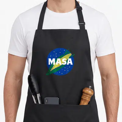 Masa mexican apron - Cartoon corn cob on blue circle with white MASA text — shown on a black cotton canvas apron against a white background