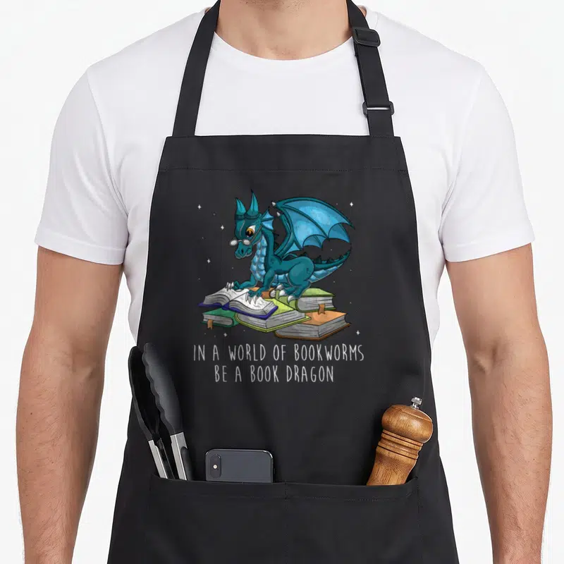 Bookworm dragon apron - Blue dragon with glasses reading books with white text IN A WORLD OF BOOKWORMS BE A BOOK DRAGON — shown on a black cotton canvas apron against a white background