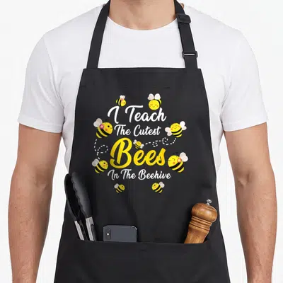 Teach bee aprons - Cartoon bees with pink flowers around stacked I Teach The Cutest Bees In The Beehive text — shown on a black cotton canvas apron against a white background
