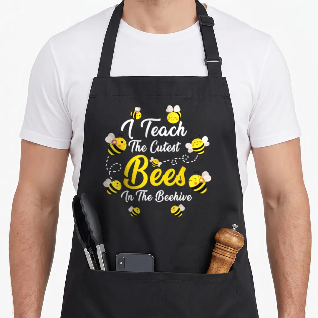 Teach bee aprons - Cartoon bees with pink flowers around stacked I Teach The Cutest Bees In The Beehive text — shown on a black cotton canvas apron against a white background